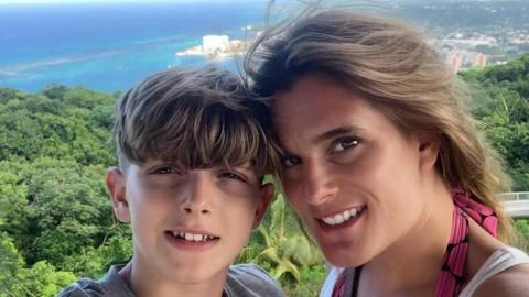 Ellen Roome with her 14-year-old son Jools Sweeney in a holiday snap. Behind them is a vibrant green forest with a very blue sea in the background. Ellen has long brown hair and brown eyes and Jools has short brown hair. They are smiling at the camera.