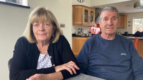 Paul and Debbie Rouget are sitting at a table in their kitchen. He is wearing a blue jumper and she is wearing a dark cardigan and she has her arms crossed.