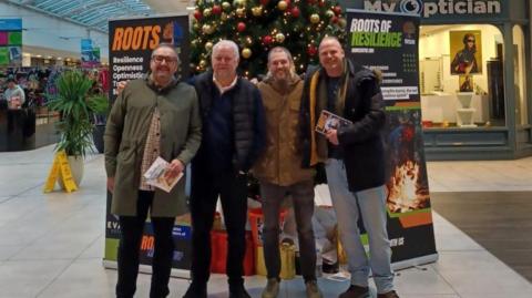 Four men standing and posing in front of a Christmas tree inside a shopping centre