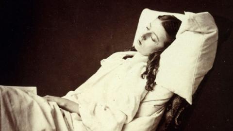 A young woman lies on a bed.. Her white clothes and pillow are in stark contrast to the dark background.