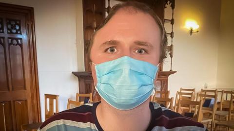 A head shot of David White looking right into the camera. He is wearing a blue hospital mask and a round-neck stripy shirt. His eye are blue and he has receding light brown hair. The right side of his head has a large indent on the top.