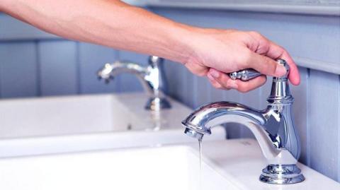 A man holding the handle of a chrome tap attached to a sink. Another chrome tap and sink are in the background. Blue tiling surrounds the taps and sinks.