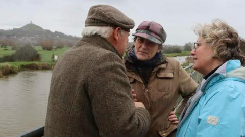 Sarah Dyke is standing between two other people by a river with Glastonbury Tor visible in the distance.