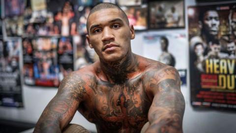 Conor Benn is shirtless and sitting in front a wall of posters