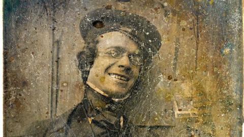 A sepia image of photography pioneer George Shaw. He is wearing Victorian clothes including a cap, shirt, jacket and neck tie. He is wearing oval glasses and smiling.