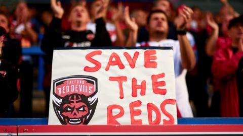 A placard with "Save The Reds" and Salford Red Devils' club crest is displayed by their fans at a recent Super League game