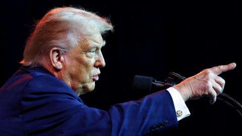 Side view of Donald Trump wearing a blue suit and a white shirt with cufflinks. He is standing before some microphones and pointing his finger.