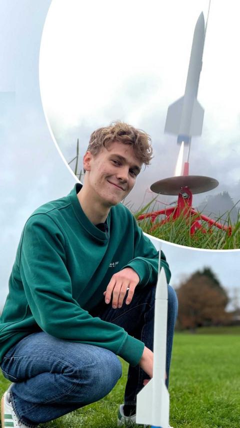 Young man stood behind a 3D-printed model rocket in a park.