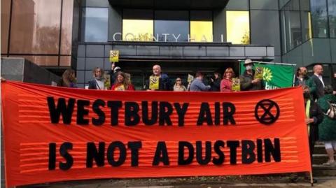 A small group of protesters stand behind a huge red banner with black lettering reading "Westbury air is not a dustbin". They are standing in front of a council building which bears a sign saying County Hall.