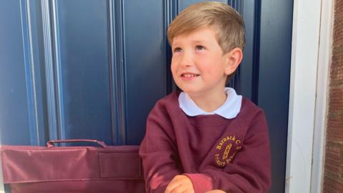 A young boy with dark blond hair is sitting on a step outside a blue door. He is wearing a burgundy school uniform with long grey socks and black shoes. The school uniform's jumper has a yellow circular logo, and he has a matching school bag next to him. He is looking up to the side and smiling.