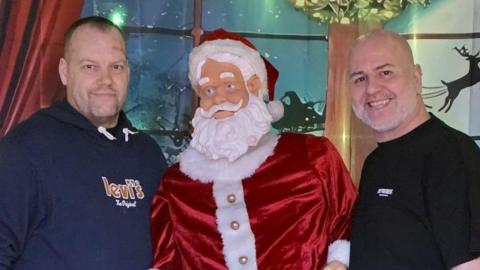 Two men standing either side of a model of Santa. The man on the left has short hair and a blue tshirt and the man on the right has a bald head and a black tshirt