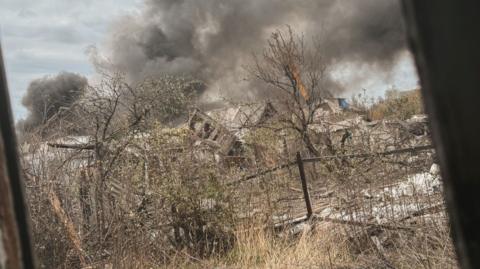 A shot through a window of a wasteland in Pokrovsk. The houses are severely damaged, the trees are bare and there is a large grey-black cloud in the distance