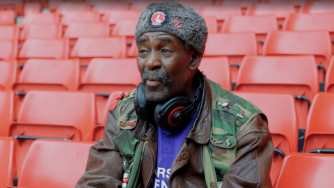 Norman Barker wearing headphones, a purple Charlton shirt, a grey hat and camouflage jacket