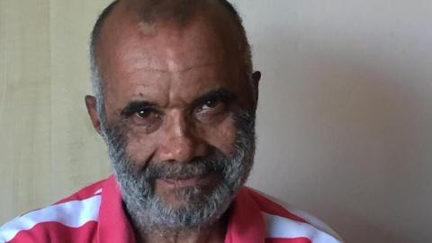 A man with a beard and short hair stands with his arms crossed, wearing a red and white striped polo shirt. He is positioned indoors against a pale wall, with a wooden door partially visible in the background. He is looking directly at the camera and has brown eyes and grey hair.
