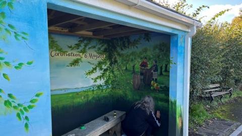 We are looking at a bus shelter which has been transformed into a beautifully decorated scene of the countryside. In the corner a grey-haired woman is adding some touches of paint to the inside wall of the bus shelter