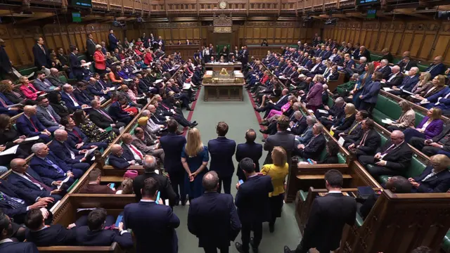 A wide shot shows MPs sat in the House of Commons. A number of MPs can be seen stood up on both sides of the chamber as they try and ask a question. Around 10 MPs are stood close to the camera as the benches are mostly full
