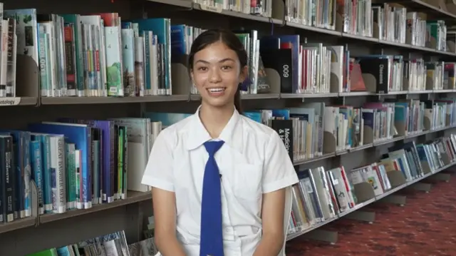 Frankie wearing her school uniform sitting in the library beside shelves of books
