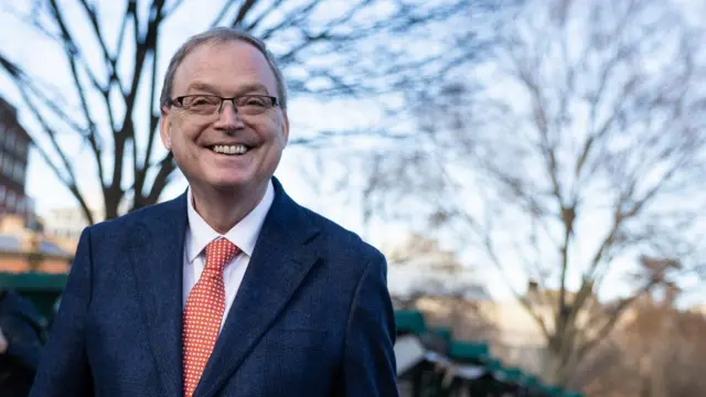 A smiling man in glasses and a blue suit and red tie with white spots smiles