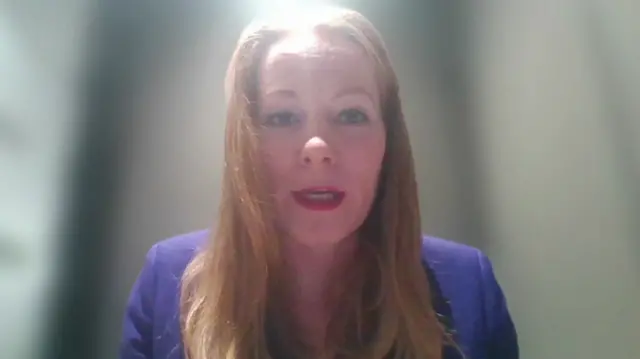 A woman with long red hair in a purple blue jacket, with a white blurred Zoom background behind her