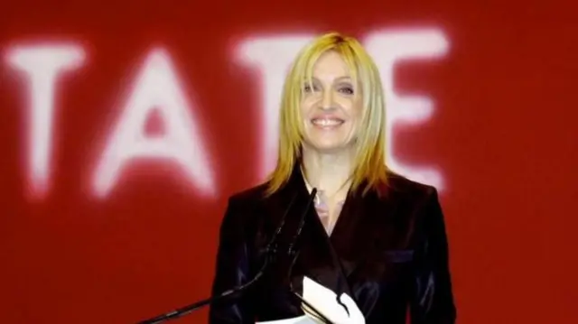 Madonna in a black jacket with straight blonde hair stands at a podium