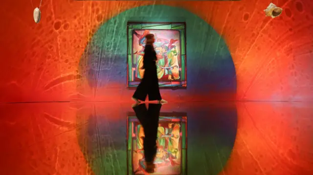 A woman walking past a large colourful painting in a gallery bathed mostly in red light and swirling shapes in Zadie Xa's installation