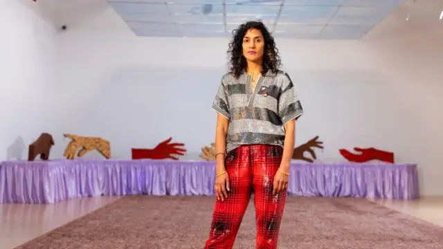 Jasleen Kaur, in red trousers, stands in front of a table with a purple cloth on it and large sculptures of hands