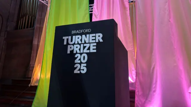 A podium with the words "Turner Prize 2025" in front of a yellow and pink curtain backdrop