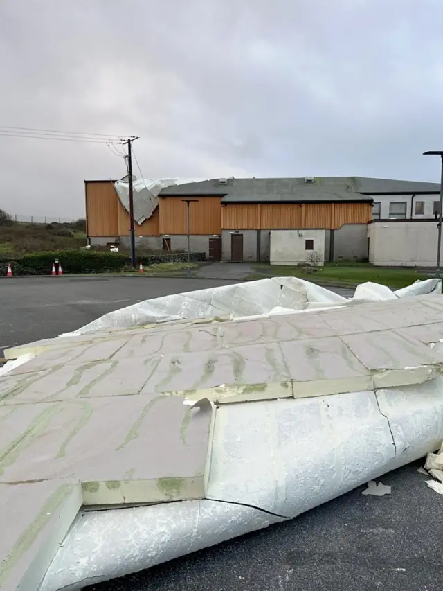 A leisure centre car park with a large piece of flat roofing on the floor. You can see the building it came from in the distance, with part of the roof drooping over it.