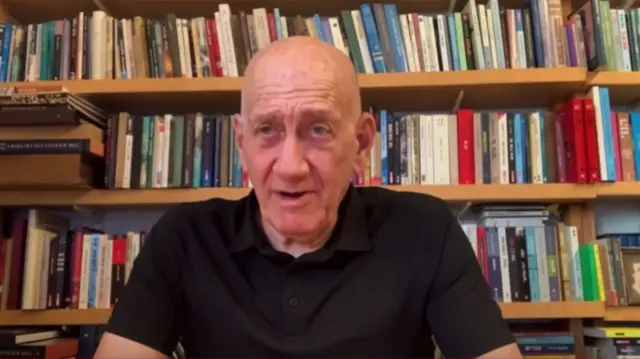 An older man with a black shirt sits in front of a bookcase.