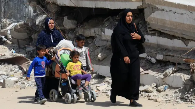 Two women and two young boys walking through street covered in rubble, a young girl sits in a pram.