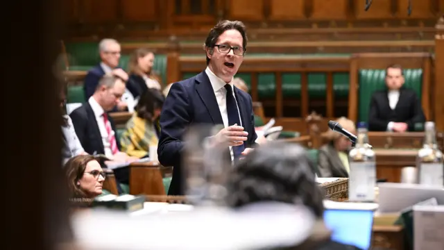 Alex Burghart wears a navy suit and tie as he speaks in the House of Commons