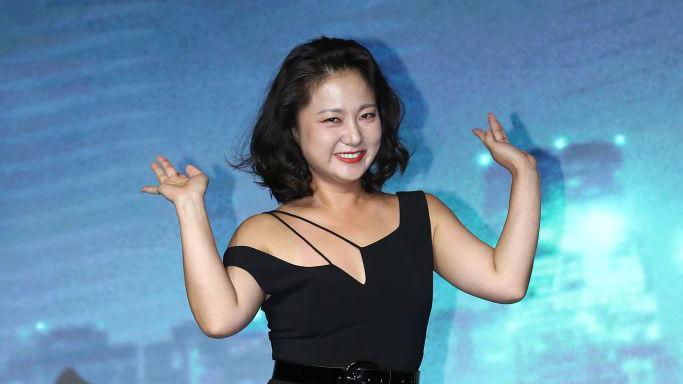 South Korean comedian Park Na-Rae with a short black bob and a black dress waving against a blue background at a press event