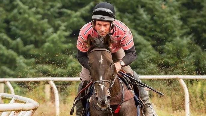 A man in a pink and black striped T-shirt with a black hat on and black boots rides a brown horse round a track with sand being thrown up in the air around them