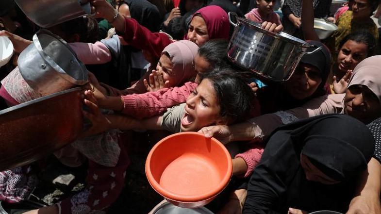 A large group of Palestinians wait to receive food cooked by a charity kitchen, in Jabalia, in the northern Gaza Strip. They are holding on pans and other receptacles and look distressed.