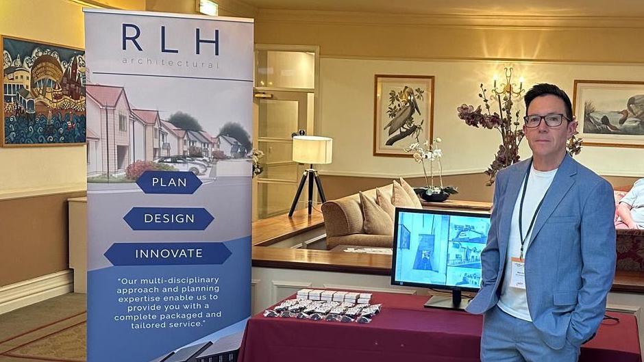 Nick Cox standing in front of a stall showcasing his company - RLH Architectural. He is wearing a blue suit jacket and trousers and a white t-shirt. He has dark hair and glasses.