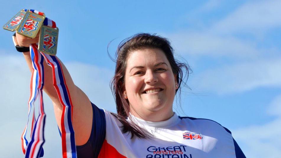 Bethan Lishman. She is wearing her Team GB kit and is holding up three medals, with red white and blue ribbons. She has brown hair, tied back but blowing in the wind, and is smiling. The sky behind her is blue with wispy clouds.