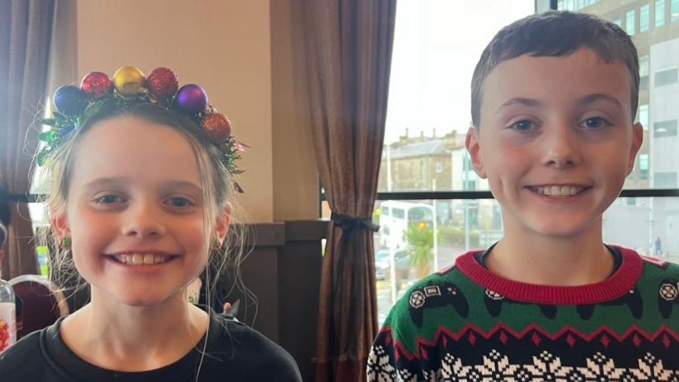 A boy and a girl, both wearing Christmas jumpers. The girl is wearing a Christmas headband.