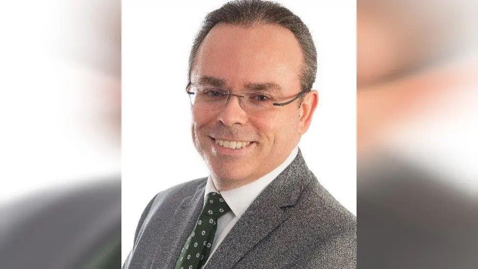 A man in a grey suit and dark green tie smiling at the camera in a portrait style photo on a white background.