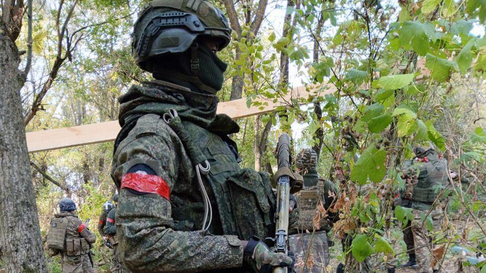 David Kuloba in full battle gear holding a gun in a forest in Ukraine with other soldiers seen in the background.