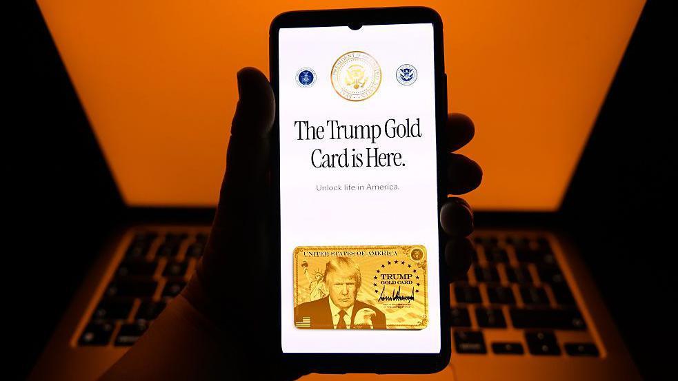 The Trump Gold Card website is displayed on a mobile phone screen posed in front of a laptop with an orange background. The Gold Card webpage says "The Trump Gold Card is Here" with the subtitle, "Unlock life in America" and an image of the card, with Trump's image
