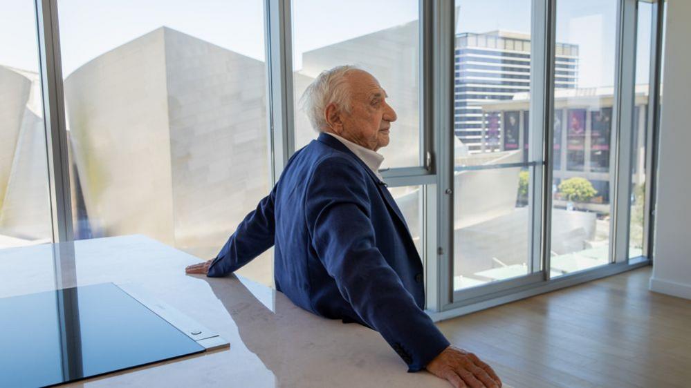 Frank Gehry has his back to the camera as he stands at a kitchen island within his personal apartment at The Grand by Gehry development in Los Angeles