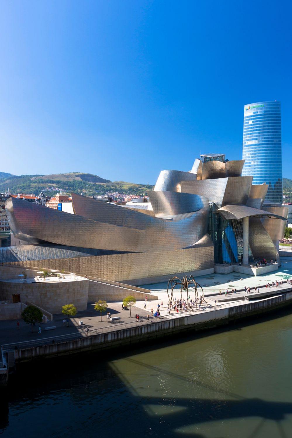 A view of the Guggenheim Museum in Bilbao, Spain. It shows the building's unique silver material, glistening in bright sunshine, around its iconic curvature.