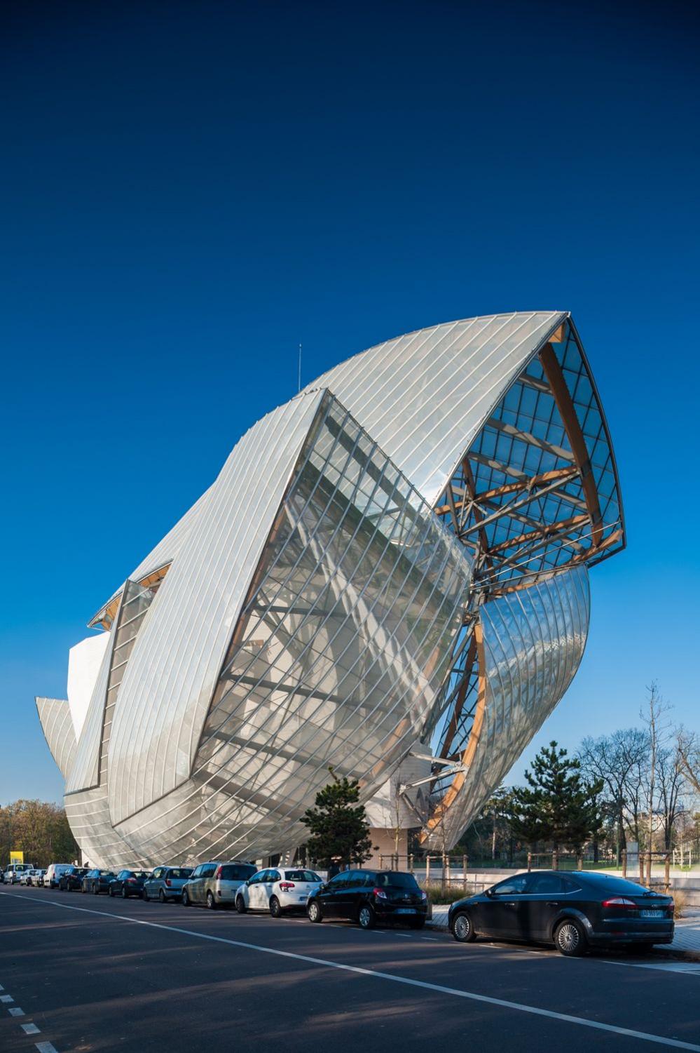 The Louis Vuitton Foundation building is seen against a bright-blue sky. Its steel and glass structure bursts out of the ground in a variety of wings surrounding a central brick building that is obscured from view by the glass. It sits beside parked cars on a road with trees in the background.