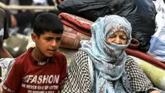 A boy and an elderly woman sit on a cart pulled by a donkey. The woman’s head is covered by a blue and white shawl.