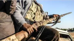 Ọwọ to gbe ibọn dani pẹlu ẹnikan to duro lẹgbẹ ẹni to gbe ibọn ọhun dani