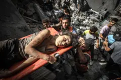 A young girl lies on a stretcher as she is pulled away from a destroyed building but emergency workers. She grips the side of the stretcher and looks disconsolate.