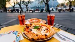 Masada pizzalar ve iki kokteyl içecek, cadde kenarındaki restoranın arka planda Como gölü manzarası var.