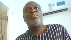 Charles Mwesigwa has short hair and greying stubble, and is wearing a navy and white striped top