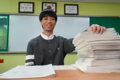 학교 책상 왼쪽엔 일반 시험지가, 오른편에는 높이 쌓인 점역 시험지가 있다. 의자에 앉아 있는 남학생이 높이 쌓인 점역 시험지에 손을 올려놓고 있다.