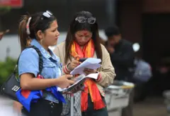 काठमाण्डू विमानस्थलमा विदेश जाने तरखरमा ��हेका दुई युवतीहरू, एक जनाले रातो टीका र खादा लगाएर मोबाइल फोन चलाइरहेकी छिन् भने अर्कीले नेपालको झण्डा भिरेर कागजपत्र लिएकी छन्